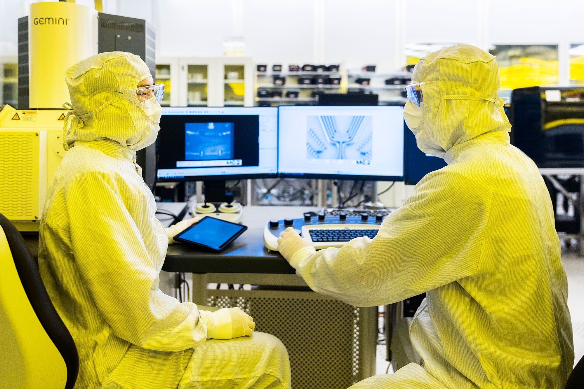Two engineers in a yellow-lit cleanroom at a scanning electron microscope; the right-hand screen shows an SEM image of a meandering device structure.