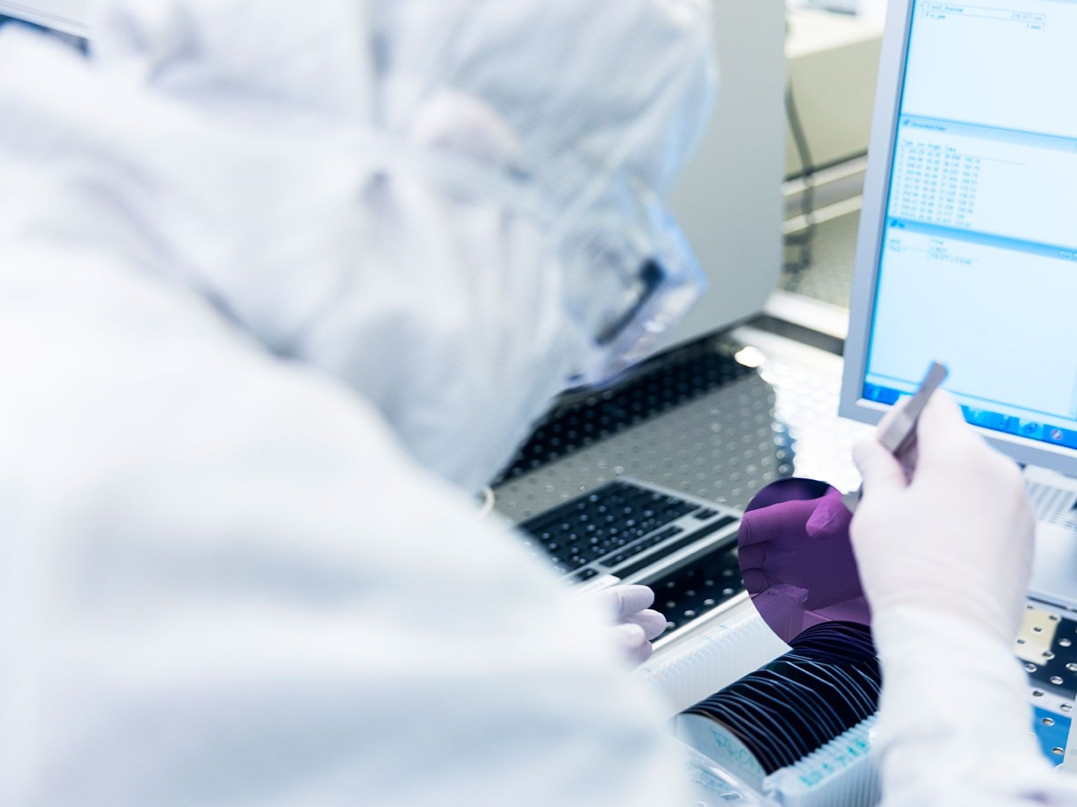 A cleanroom engineer in a white suit and hood lifts a wafer from a cassette with tweezers, next to a measurement screen showing fitted curves.