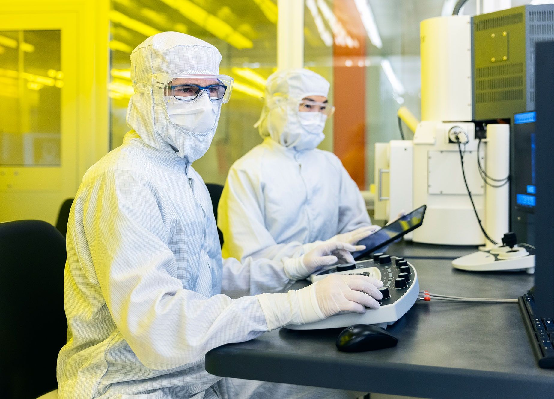 Two engineers in cleanroom suits, hoods and masks work at an electron-beam workstation under yellow lithography light.