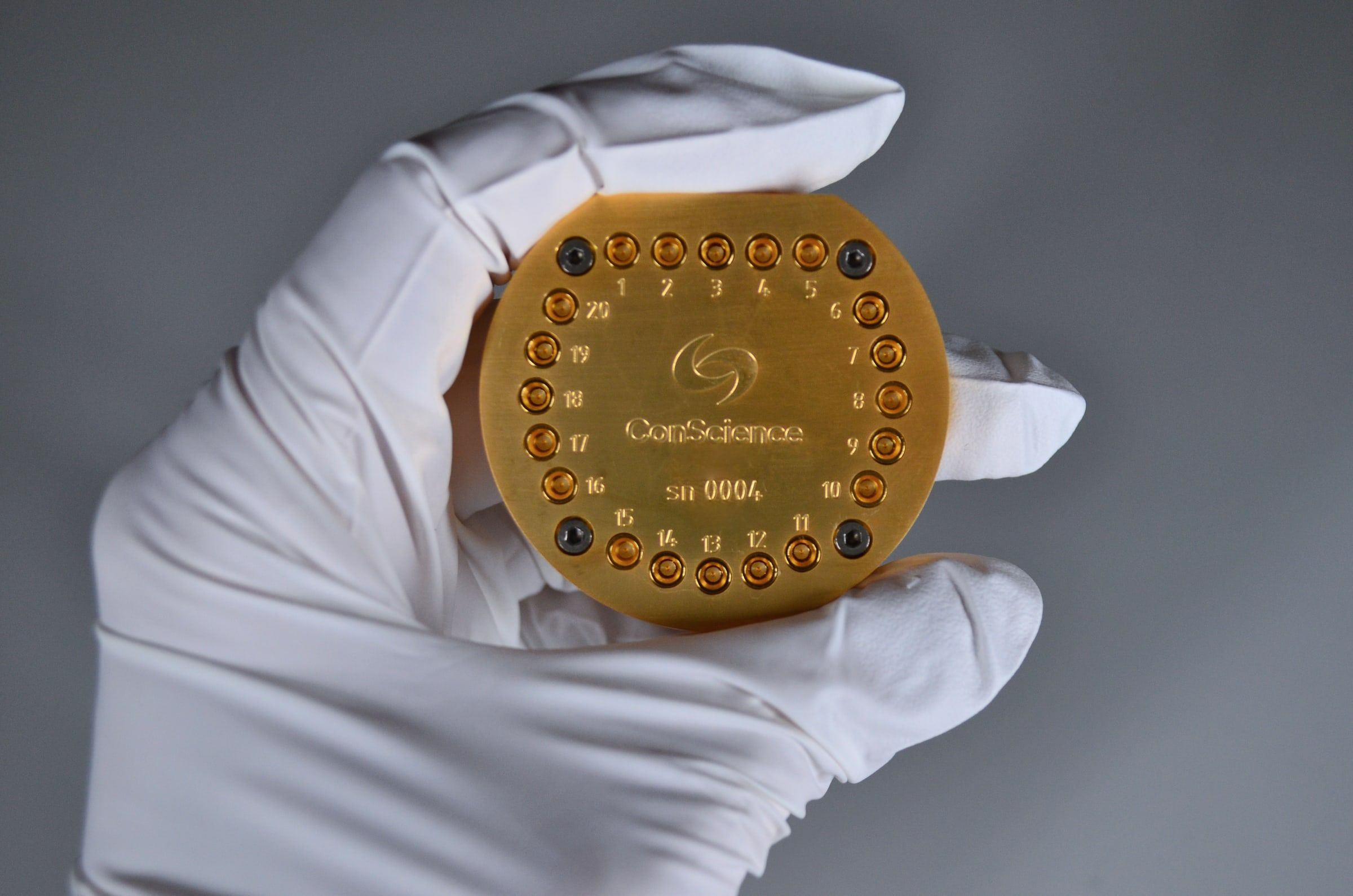 A gloved hand holds the QiB2 in its Box20 package: a round gold-plated box with ports numbered 1 to 20 around the rim and the ConScience logo engraved on top.